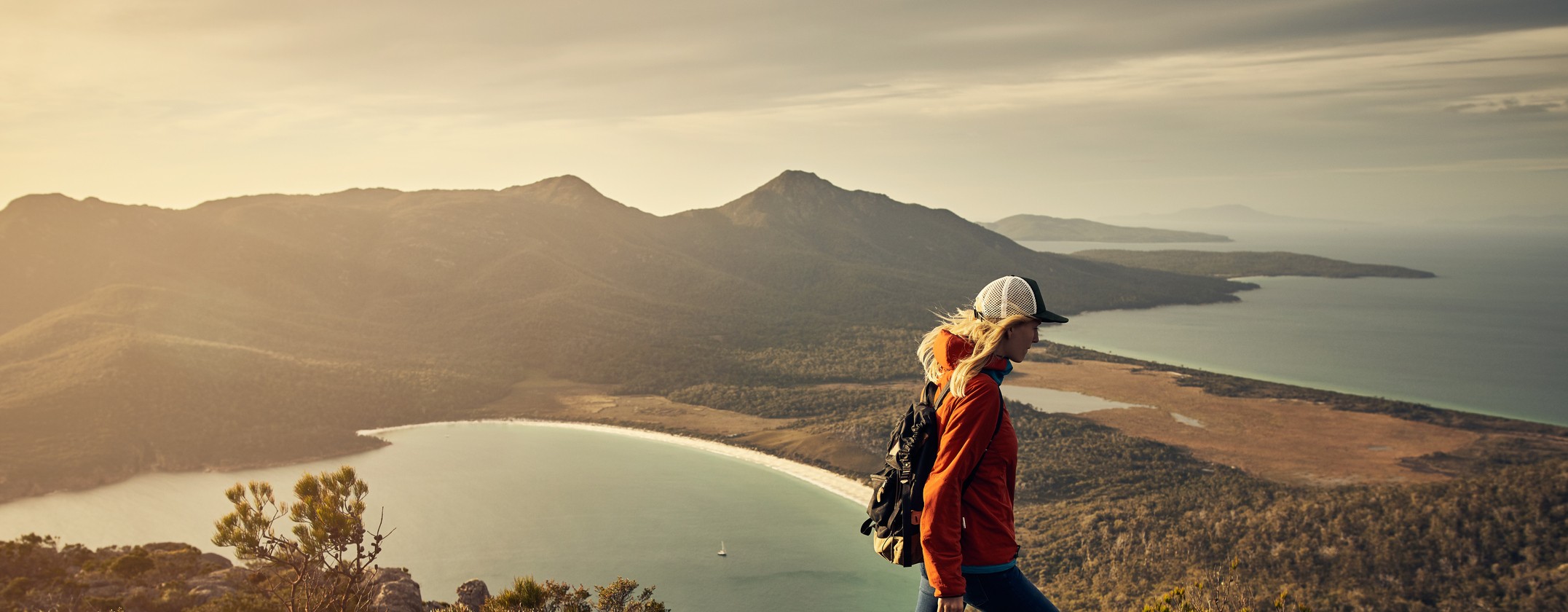 A woman hikes a hill with a beach and bay below her in the distance.